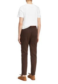 Osoba ubrana w białą koszulę z krótkim rękawem, ciemnobrązowe spodnie z tylnymi kieszeniami oraz jasnobrązowe, sznurowane buty casual, stojąca tyłem.