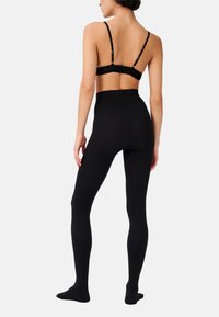 Leggings neri con vita alta, texture liscia, design a lunghezza intera e materiale morbido, indossati con un bralette nero coordinato.