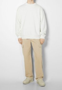 Weißer Sweatshirt mit gerippten Bündchen, kombiniert mit beigen Cargo-Shorts mit Seitentaschen und Ziernähten, abgerundet mit beigen Sneakers.