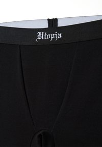 Prenda de tela negra con estrías y una amplia cinturilla negra que presenta letras blancas con la palabra "Utopia". El diseño incluye una abertura funcional.