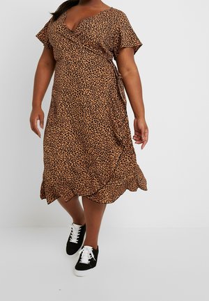 Kvinde iført en brun leopardprint slå-om-kjole med korte ærmer og sorte sneakers med hvide snørebånd, stående mod en ensfarvet baggrund.