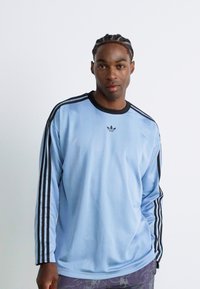 Young man with braided hair wearing a light blue Adidas long-sleeve shirt with black stripes and logo, standing against a plain white background.