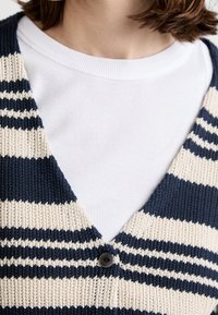 Striped navy and cream knitted cardigan with a v-neck, featuring a single black button. Worn over a white ribbed crew neck shirt.