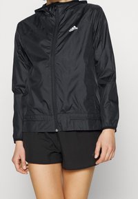 Donna che indossa una giacca a zip con cappuccio nera Adidas e pantaloni sportivi neri, in piedi contro uno sfondo uniforme.