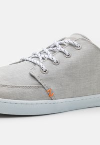 Helle graue Stoffschuhe mit weißen Schnürsenkeln, ausgestattet mit silbernen Ösen und einem orangefarbenen gestickten Logo. Gummisohle mit strukturiertem Griffmuster.