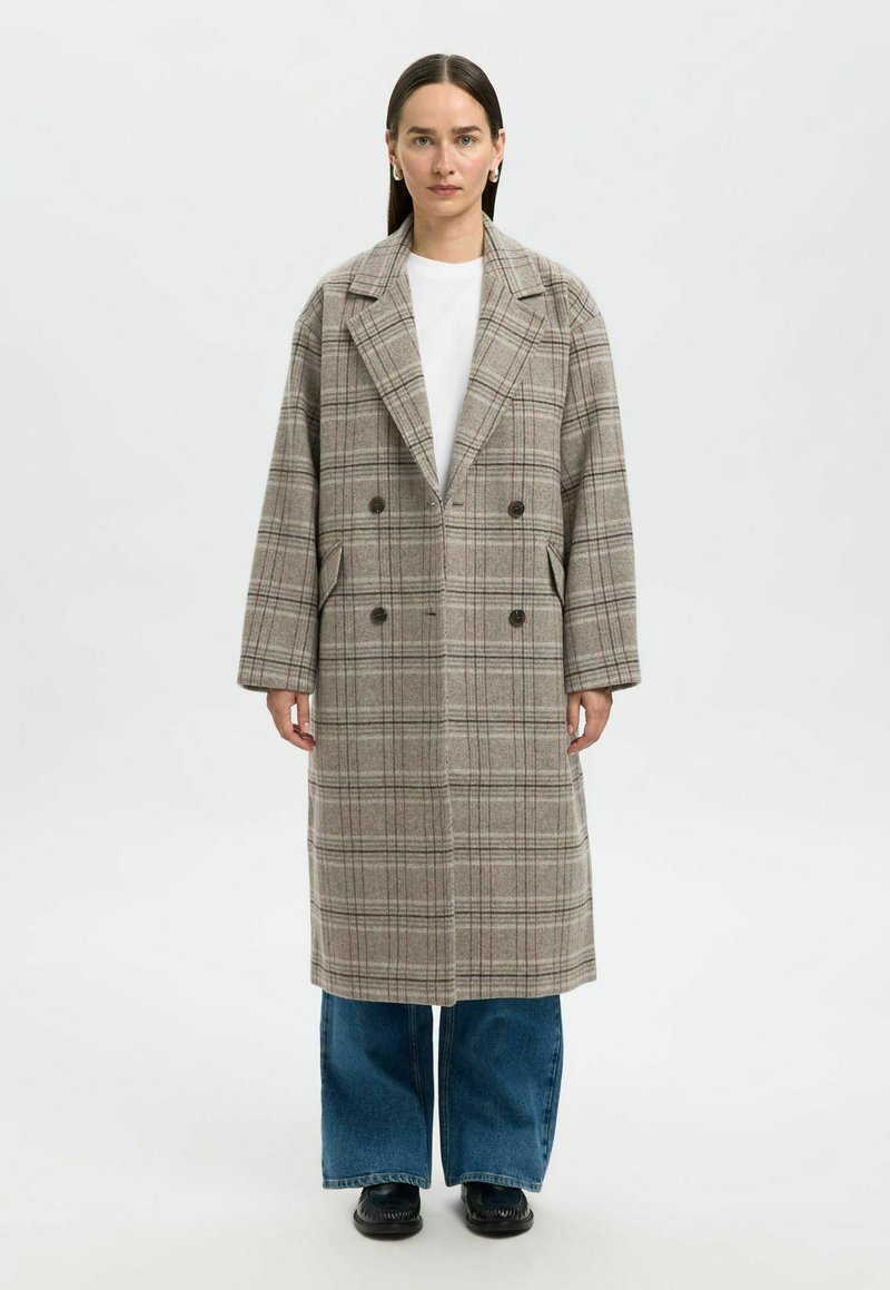 Femme aux cheveux raides et foncés portant un long manteau beige à carreaux, une chemise blanche, un jean bleu à jambes larges et des chaussures noires, debout devant un fond blanc.