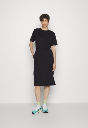 Vestido preto de t-shirt até ao joelho, com mangas curtas, cintura cingida e bolsos laterais. O modelo usa ténis multicoloridos com meias verdes.
