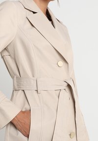 Beige trenchcoat med en dubbelknäppt design, stora guldknappar, midjebälte och sidofickor, med ett slätt, strukturerat tyg.