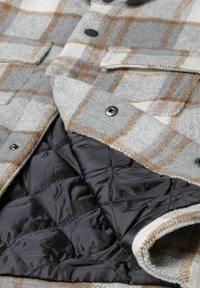 Veste à carreaux gris et blanc avec des accents marron, dotée de fermetures à boutons et d'une doublure matelassée noire. Deux poches avant sont visibles.