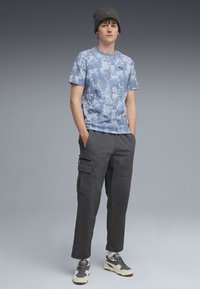 Blaue T-Shirts mit abstraktem Muster, schwarze Cargo-Hosen mit Taschen, graue Mütze und grau-weiße Sneakers. Neutraler Hintergrund.