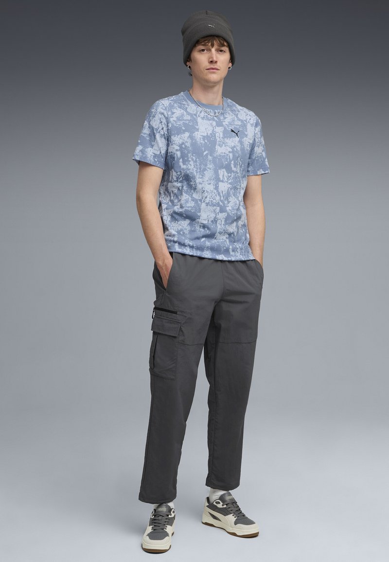 Blaue T-Shirts mit abstraktem Muster, schwarze Cargo-Hosen mit Taschen, graue Mütze und grau-weiße Sneakers. Neutraler Hintergrund.