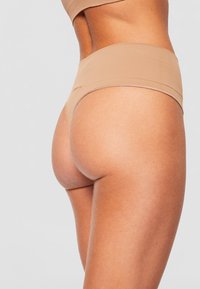 Beige, hoch taillierter Tanga mit minimaler Bedeckung, glatter Textur und elastischem Bund. Nahtloses Design, das für Komfort ausgelegt ist.