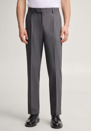 Homme portant un pantalon de costume gris avec pli central, des chaussures habillées noires et une chemise blanche, visible de la taille aux chaussures sur fond uni.