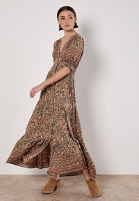 Robe maxi dans des tons terreux avec un motif floral en paisley, présentant un décolleté en V profond et de courtes manches bouffantes. Des sabots marron sont portés.