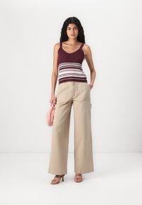 Débardeur bordeaux en tricot avec des rayures blanches, associé à un pantalon large beige. Le modèle tient une pochette rose. Sandales à lanières.