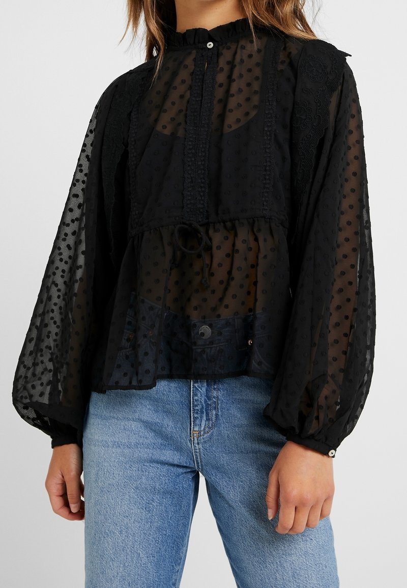 Femme portant un chemisier noir transparent à pois, avec des détails en dentelle et des manches longues bouffantes, associé à un jean bleu.