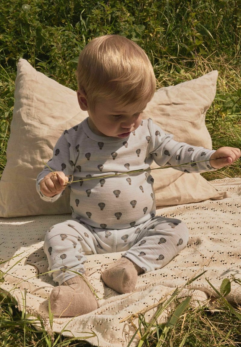 Bébé assis sur une couverture à l'extérieur, portant un pyjama avec des imprimés de montgolfières, tenant et étirant un brin d'herbe fin.