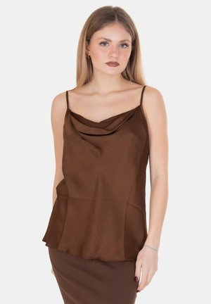 Giovane donna con capelli biondi lisci che indossa un top marrone in raso senza maniche e gonna abbinata, in piedi davanti a uno sfondo bianco semplice.