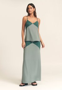 Lysegrøn maxikjole med spaghetti-stropper, lagdelt design med kontrasterende nuancer og glat tekstur. Bæres med sorte flade sandaler.