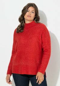Roter Strickpullover mit hohem Kragen, geripptem Saum und seitlichen Schlitzen. Weicher Stoff, lange Ärmel und lässige Passform, kombiniert mit dunklen Jeans.