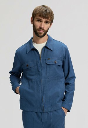 Uomo con barba che indossa una giacca blu con cerniera e tasche sul petto con bottoni, abbinata a pantaloni blu, in piedi con le mani nelle tasche.