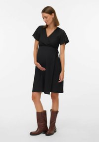 Robe portefeuille noire avec manches courtes évasées, dotée d'une ceinture à la taille et d'un ourlet au niveau des genoux. Portée avec des bottes marron montant jusqu'au genou.