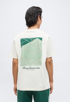 Homme portant un t-shirt blanc avec un graphique abstrait de terrain de football vert et le texte "Scandinavian Football Community" dans le dos.