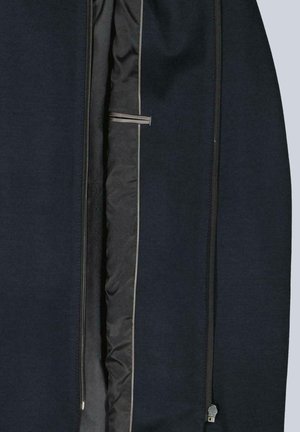 Innenansicht einer schwarzen Jacke mit gepolstertem Innenfutter, Innentasche mit Einfassung und zwei vertikalen Reißverschlüssen vor grauem Hintergrund.