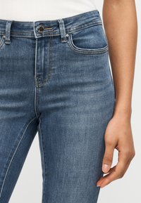 Κοντινό πλάνο ατόμου που φοράει μπλε τζιν τζιν με κουμπί και μπροστινές τσέπες, το χέρι χαλαρό στο πλάι, πάνω σε φωτεινό φόντο.