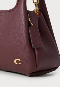 Burgundy leather handbag with a textured finish, gold hardware, and a small tag featuring the brand logo. Rounded shape with a seam detail.