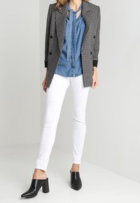 Blazer doppiopetto a zig zag bianco e nero sopra una camicia di jeans blu, abbinato a jeans skinny bianchi e mule con tacco blocco neri.