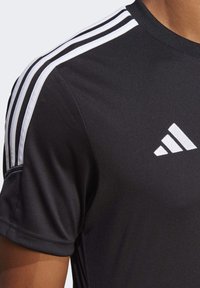 Svart idrottströja med korta ärmar, med tre vita ränder på axlarna och en vit logotyp. Slät, fukttransporterande tyg.