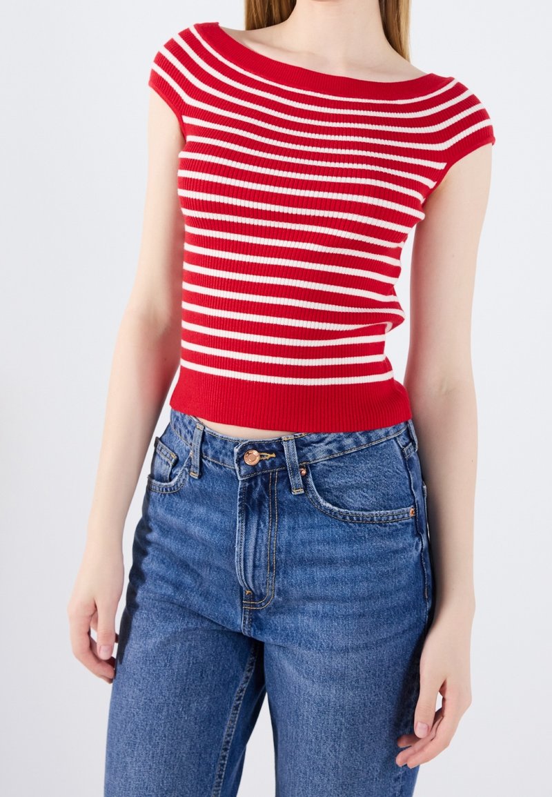 Femme portant un haut sans manches à rayures horizontales rouges et blanches et un jean bleu taille haute, debout devant un fond uni.