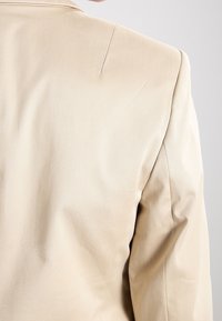 Blazer beige su misura con un tessuto liscio, spalle strutturate e una texture sottile. La vista posteriore mostra linee pulite e dettagli minimi.