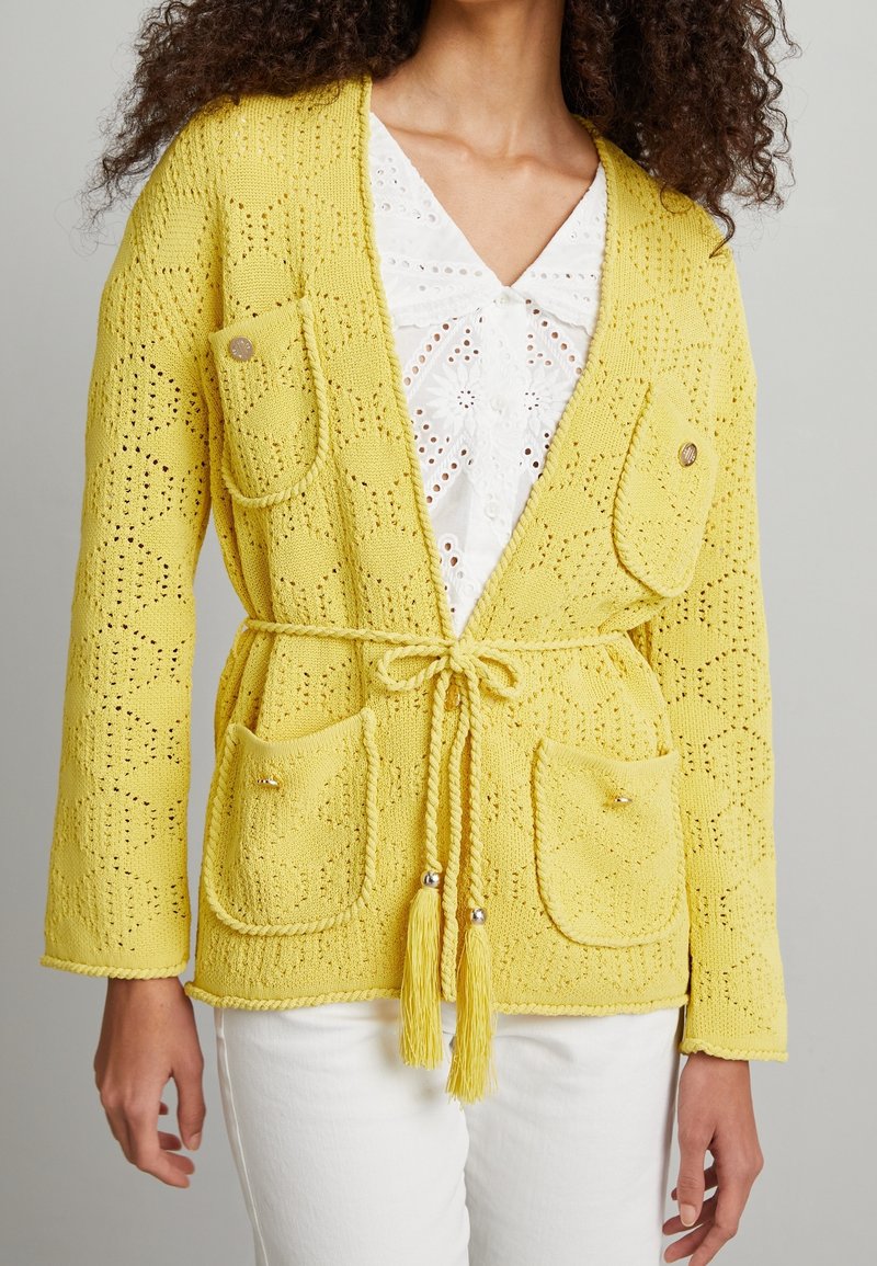 Kobieta ubrana w żółty dzianinowy kardigan z fakturowanym wzorem, przednimi kieszeniami i paskiem z frędzlami, na białej bluzce z ażurowym wzorem i białych spodniach.