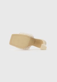 Heeled sandal with a square shape, made of beige material. Features a textured sole and embellished with white pearl accents on the upper.