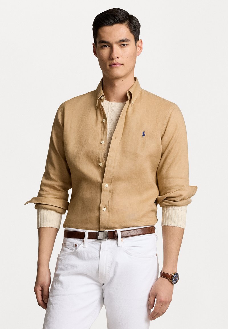 Chemise en lin marron clair, boutonnée, avec des manches retroussées. Portée par-dessus un pull crème, associée à un jean blanc et une ceinture marron.