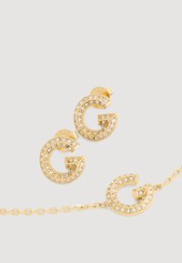 Set zlatnog nakita koji uključuje okrugle naušnice i narukvicu s 'G' dizajnom, ukrašenu sjajnim prozirnim rhinestones za dodatni sjaj.