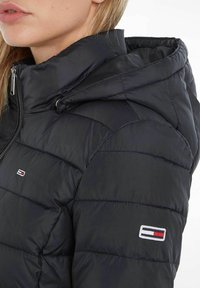 Black puffer jacket with a zippered hood, quilted design, and embroidered logo on the sleeve. Smooth texture and fitted shape.