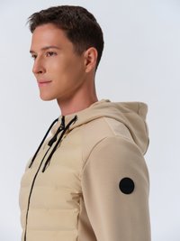 Beige pufferjack met een gladde stofstructuur, voorzien van een capuchon, een zwarte rits en een rond zwart logoaccent op de mouw.