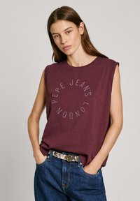 Burgundská bezrukávová tričko vyrobená z měkké látky, s kulatým potiskem "PEPE JEANS LONDON" zdobeným třpytivými kamínky, v kombinaci s modrými džíny.