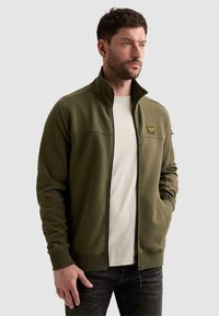 Homme aux cheveux courts et bruns, avec une barbe, porte une veste zippée texturée vert olive et une chemise blanche, debout, une main dans la poche.