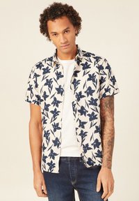 Lyhythihainen nappipaita beigen sävyssä, jossa on tummansiniset kukkakuviot. Ominaisuuksiin kuuluu rento istuvuus ja yksinkertainen valkoinen aluspaita, joka on yhdistetty farkkuihin.