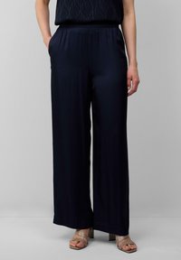 Pantalons larges en marine foncé, fabriqués à partir d'un tissu lisse. Présente une taille élastique et des poches latérales. Associés à des sandales à talons blocs beiges.
