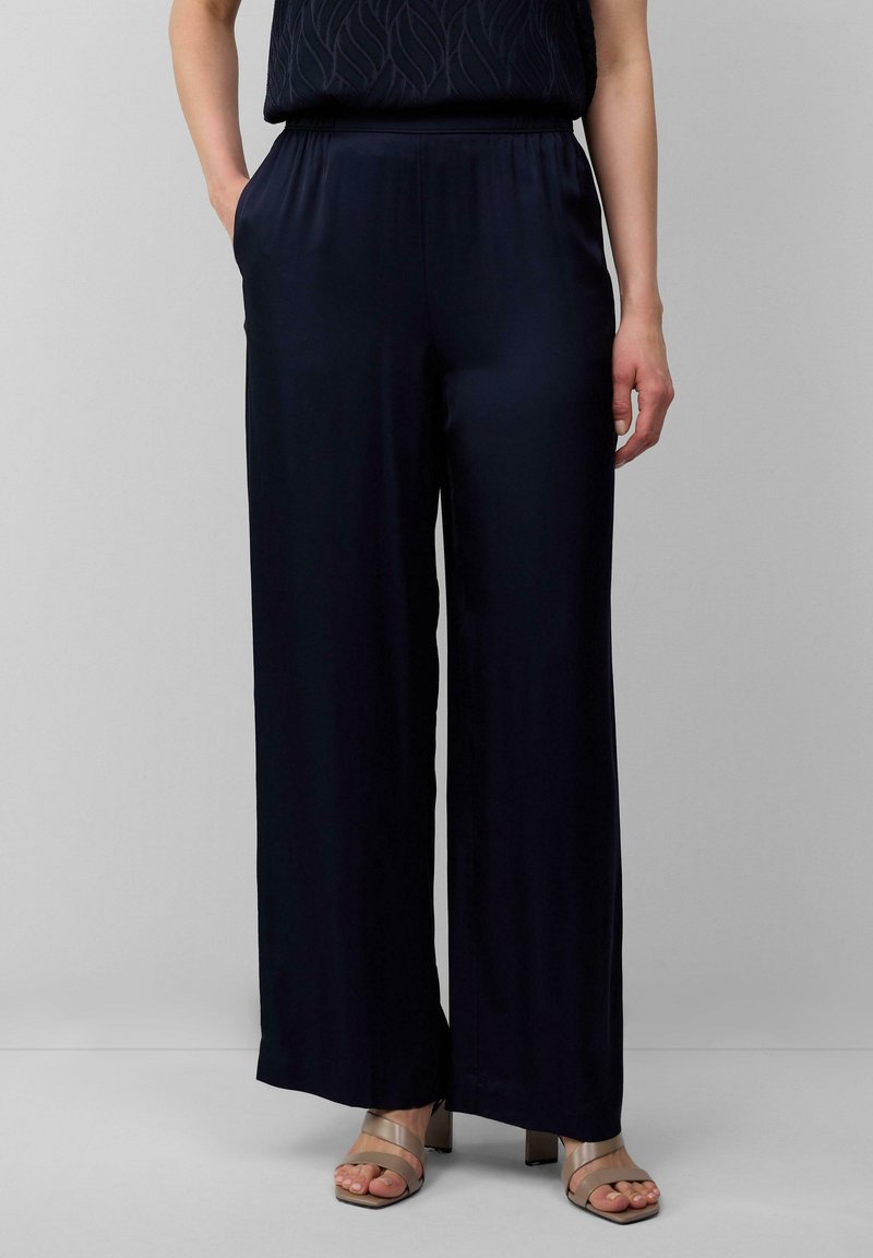 Pantalons larges en marine foncé, fabriqués à partir d'un tissu lisse. Présente une taille élastique et des poches latérales. Associés à des sandales à talons blocs beiges.