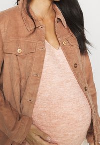 Veste en velours côtelé orange rouille avec des boutons dorés, superposée à un pull texturé rose clair, mettant en valeur un ventre arrondi.