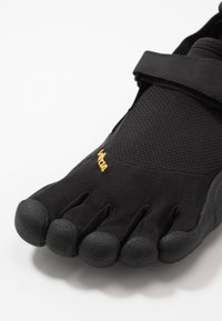 Chaussure noire à cinq orteils avec une tige en mesh, une sangle velcro et une semelle en caoutchouc texturé. Présente un design minimaliste et un logo jaune audacieux.