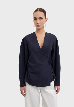 Vrouw met naar achteren getrokken haar, wit ovale oorbellen, een marineblauwe overslagblouse gestrikt in de taille en witte broek tegen een effen achtergrond.