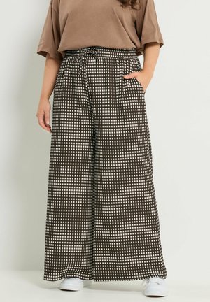 Kvinde iført vide sort-hvide ternede bukser med snøre i taljen, tan farvet løs skjorte og hvide sneakers, stående med den ene hånd i lommen.