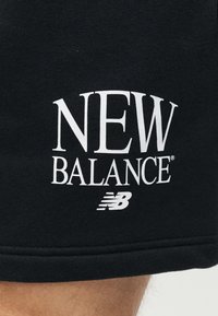 Κοντινό πλάνο μαύρου υφάσματος με λευκό κείμενο "NEW BALANCE" και λογότυπο τυπωμένα κοντά στο τελείωμα πάνω από γυμνό πόδι.